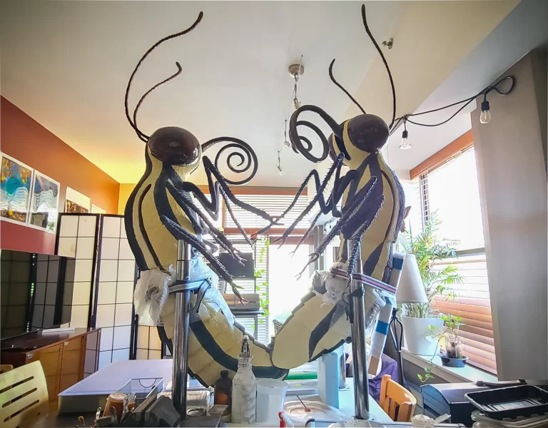 Sculptural installation in residential space featuring two suspended pale forms with segmented bodies, black striping, and curved spiral appendages positioned in interactive pose. Large-scale creatures with bulbous heads transform domestic interior into otherworldly environment.