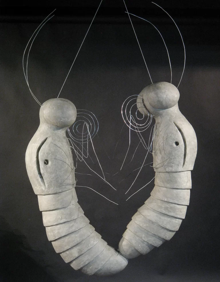 Sculptural installation featuring two suspended pale segmented forms with bulbous heads positioned in interactive pose against dark background. Linear wire elements create spiral patterns suggesting metamorphic transformation and communication between stylized larval forms.
