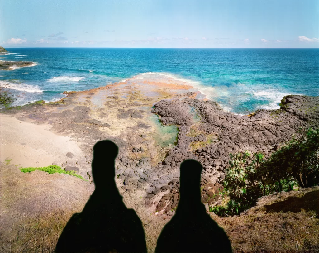 In shadow dodo reconstructions positioned on coastal bluffs at Le Gris Gris, Mauritius, gazing toward turquoise sea and lava rock structures. The outlined vanished species artworks form a poignant display within their ancestral seaside habitat.