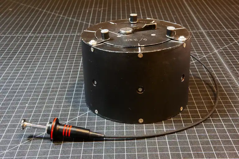 Handcrafted panoramic 6x17cm 250° multi-pinhole camera built from a modified Mamiya RB film back. The matte black cylindrical housing features multiple drilled pinholes and a top-mounted winding lever marked with exposure rotation indicators. A black and red cable release is attached, and the camera is placed on a grid-lined work surface.
